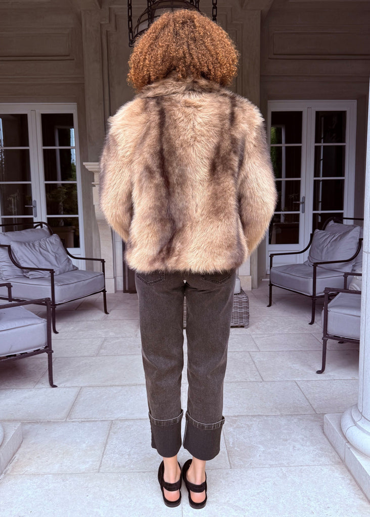 Person wearing a fur coat standing on a patio with white columns and chairs.