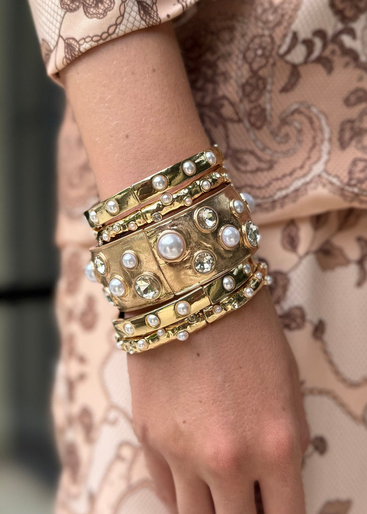 Close-up of a hand wearing multiple gold bracelets with pearls against a patterned garment.