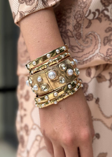 Close-up of a hand wearing multiple gold bracelets with pearls against a patterned garment.