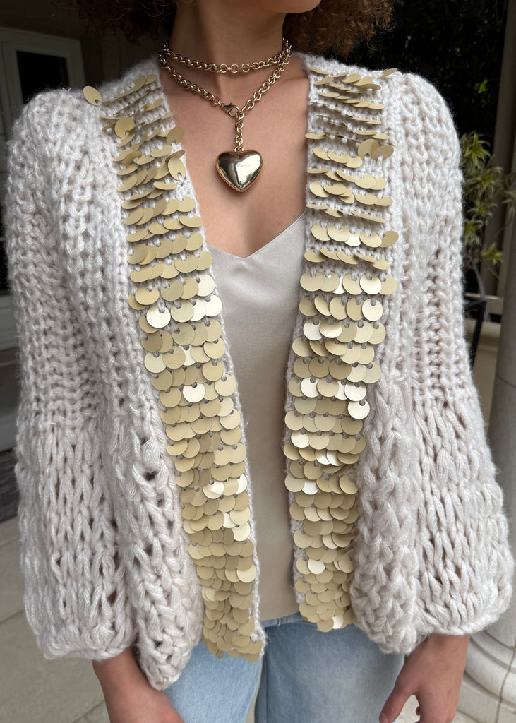 Person wearing a white knit cardigan with gold sequin details, standing indoors.