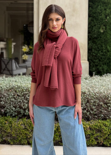 Woman wearing a pink sweater and scarf standing in front of a building with greenery.