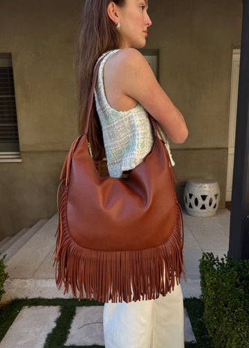 Woman carrying a brown leather handbag with fringe in an outdoor setting.