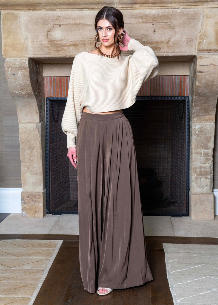 Person wearing a beige sweater and brown wide-leg pants standing in front of a fireplace.