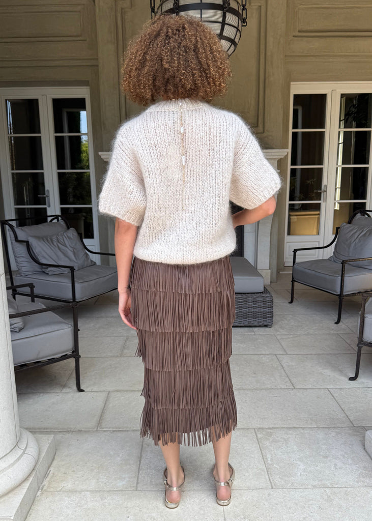 Person wearing a white top and brown fringed skirt standing in an outdoor setting with chairs and a building in the background.