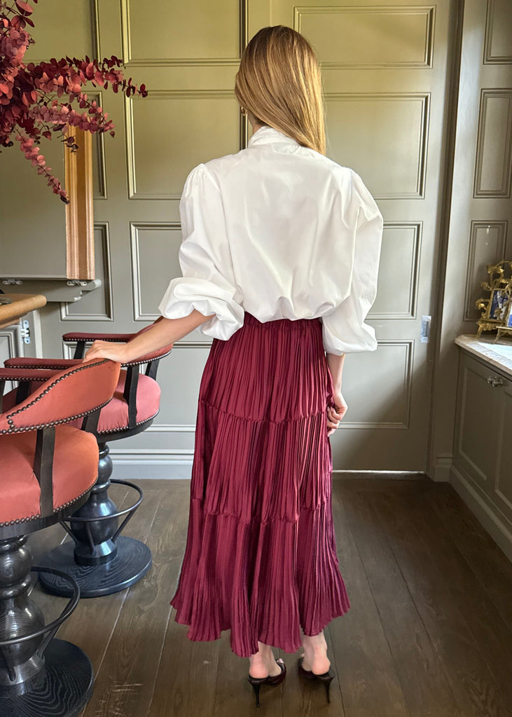 Woman in a white blouse and burgundy skirt standing in a room with wooden flooring and paneling.