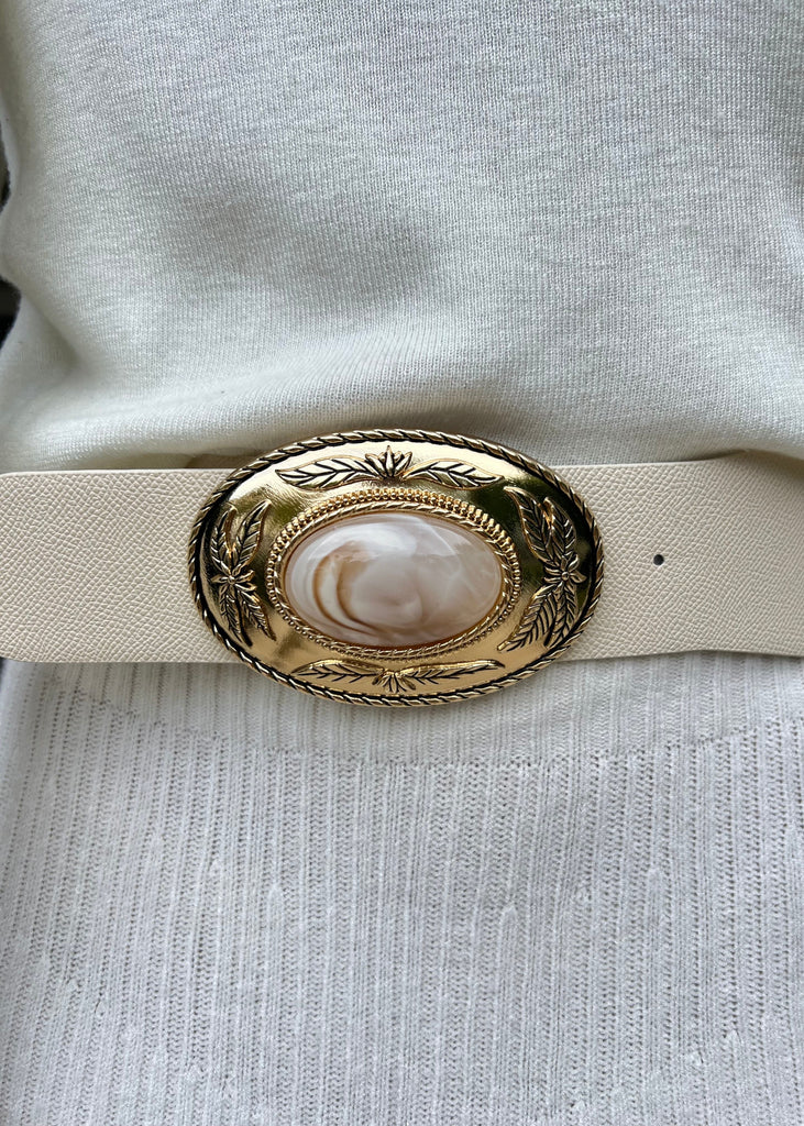Beige belt with a decorative gold buckle featuring a central pearl-like stone, worn on a light gray outfit.