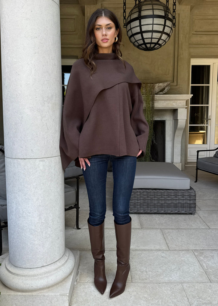 Woman in a brown sweater and jeans standing in an outdoor setting with columns and furniture.
