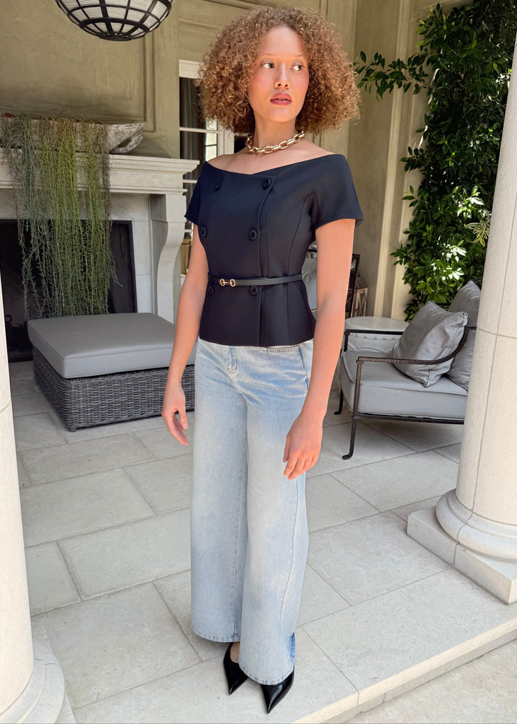 Woman in a black top and light blue jeans standing in an outdoor setting with columns and plants.