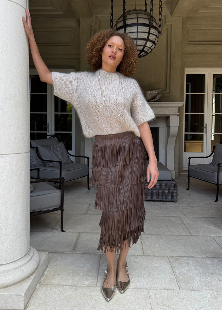 Woman standing in a stylish outdoor setting wearing a white top and brown fringed skirt.