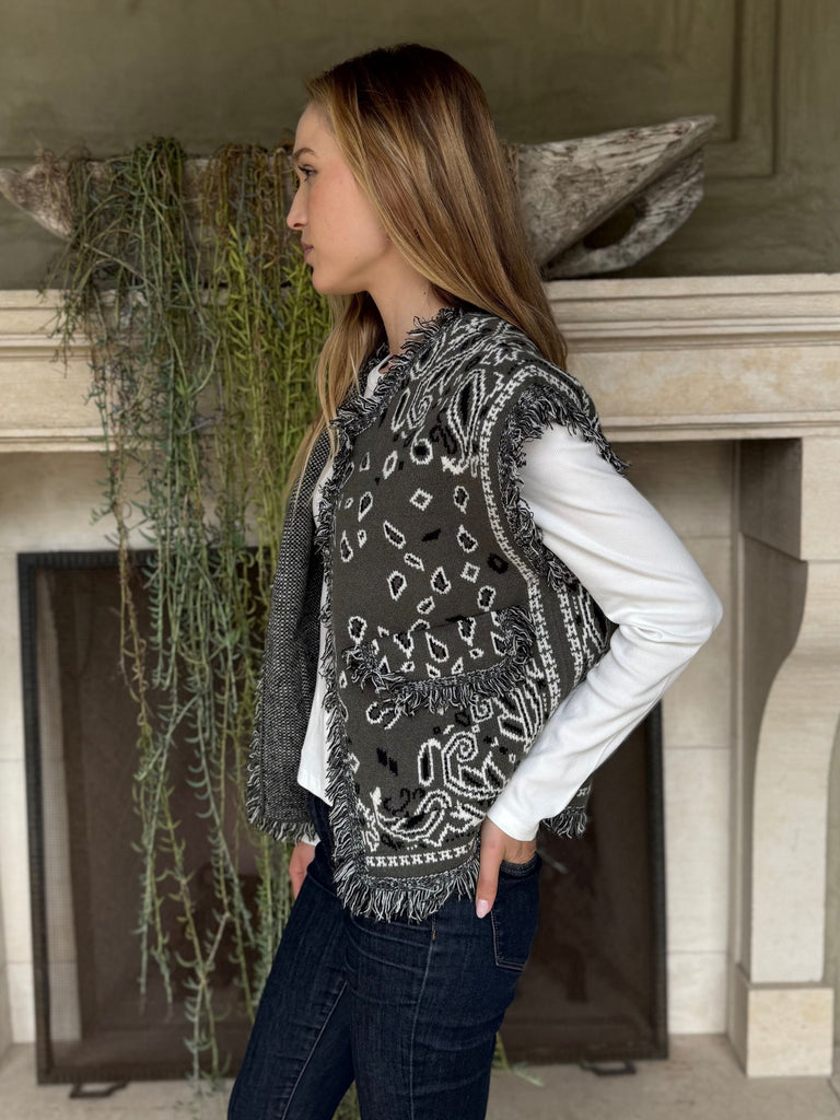 Woman wearing a patterned vest over a white shirt in front of a fireplace.