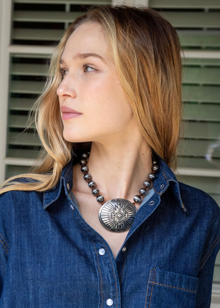 Woman wearing a blue denim shirt with a necklace, standing indoors.