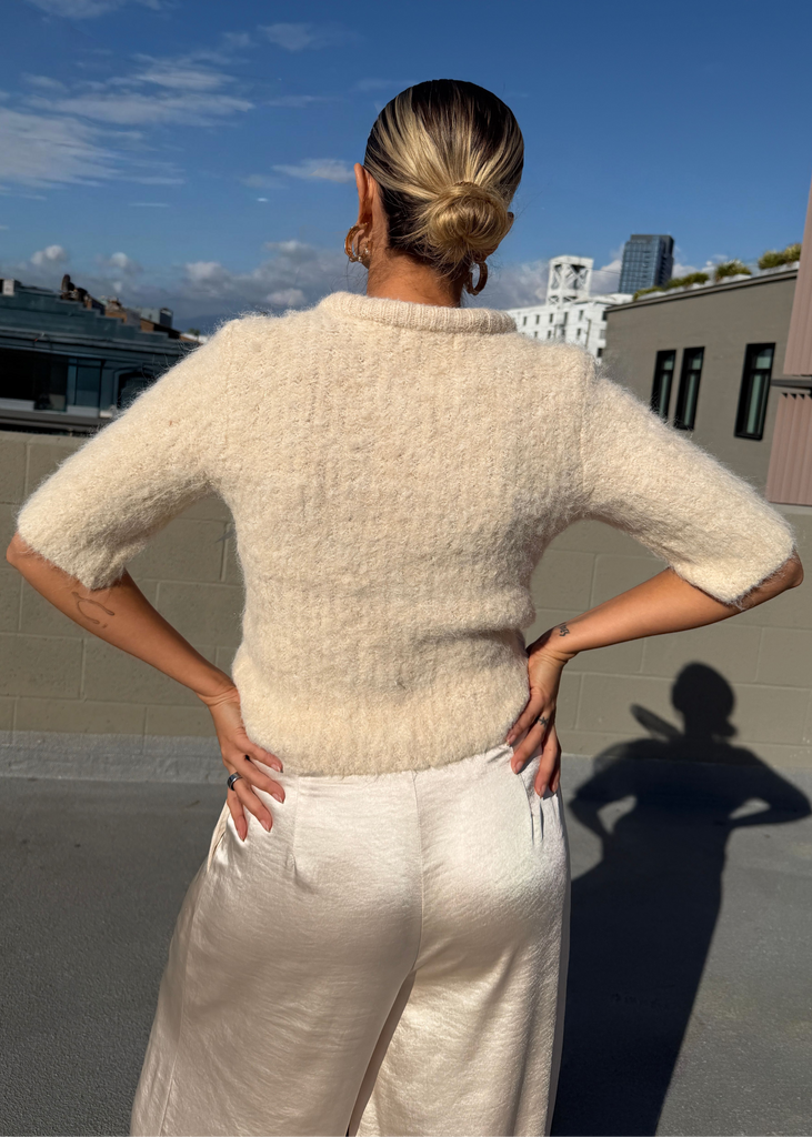 Person wearing a light-colored sweater and white pants on a rooftop with a clear blue sky.