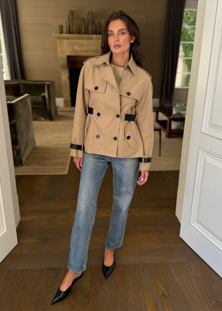Woman wearing a beige trench coat and blue jeans standing in a room with wooden flooring and white walls.