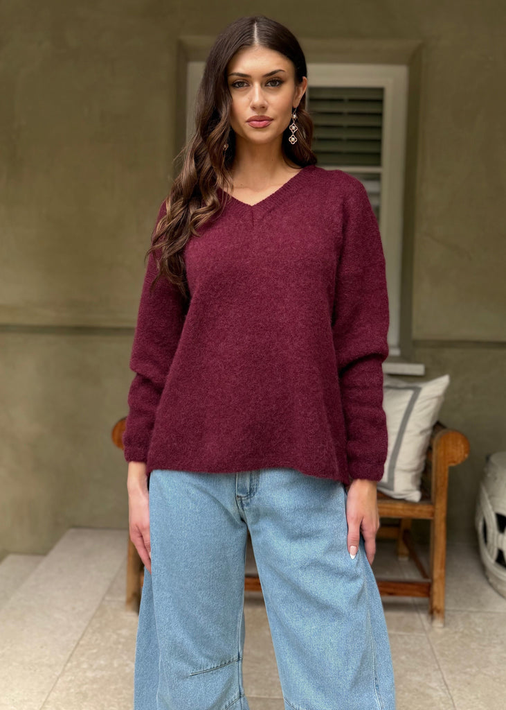 Woman wearing a maroon sweater and light blue jeans standing in front of a beige wall.