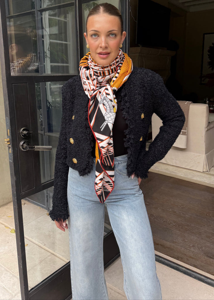 Woman wearing a black jacket with gold buttons, light blue jeans, and a patterned scarf standing in front of a glass door.