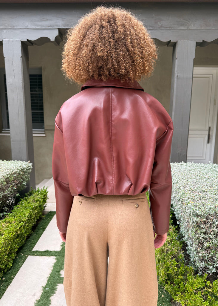 Person wearing a pink leather jacket and beige pants standing in a garden.