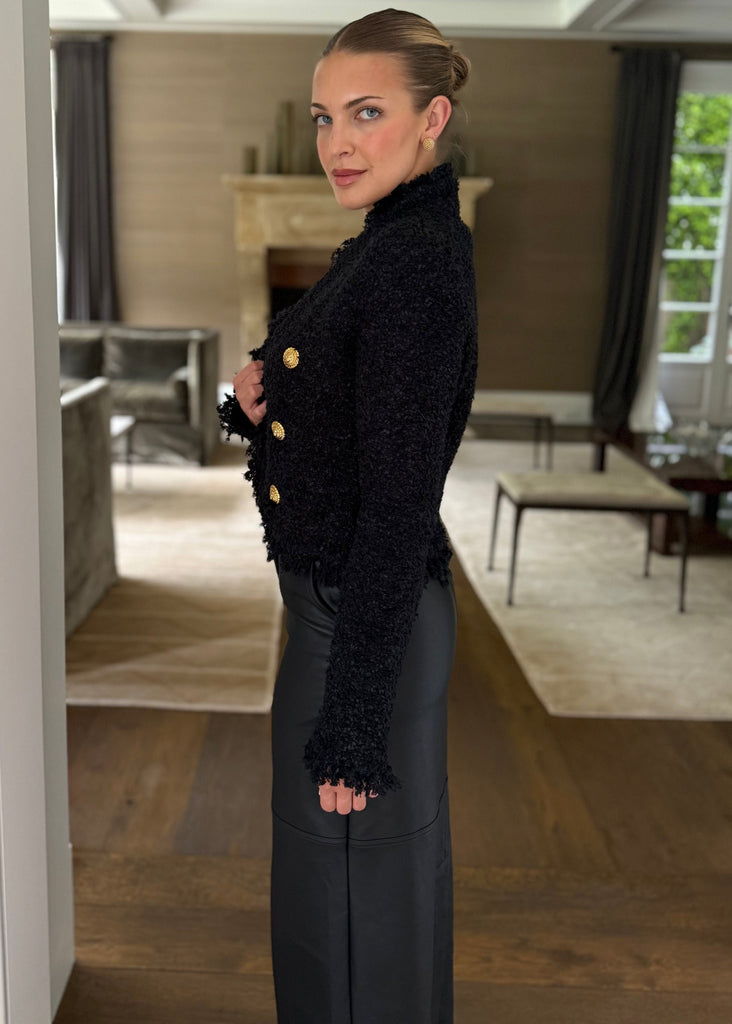 Woman in a black outfit with gold buttons standing in a room with wooden flooring and a large mirror.