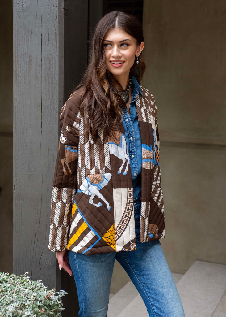 Woman wearing a patterned jacket and blue jeans standing against a neutral wall.