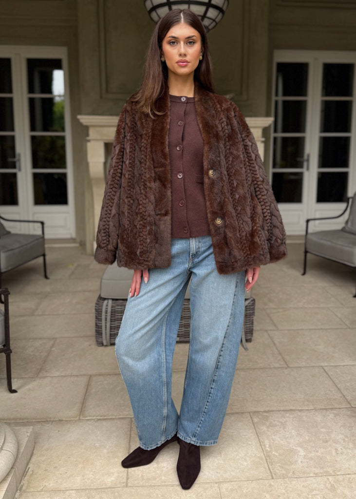 Woman wearing a brown fur coat, brown top, and blue jeans standing on a patio.