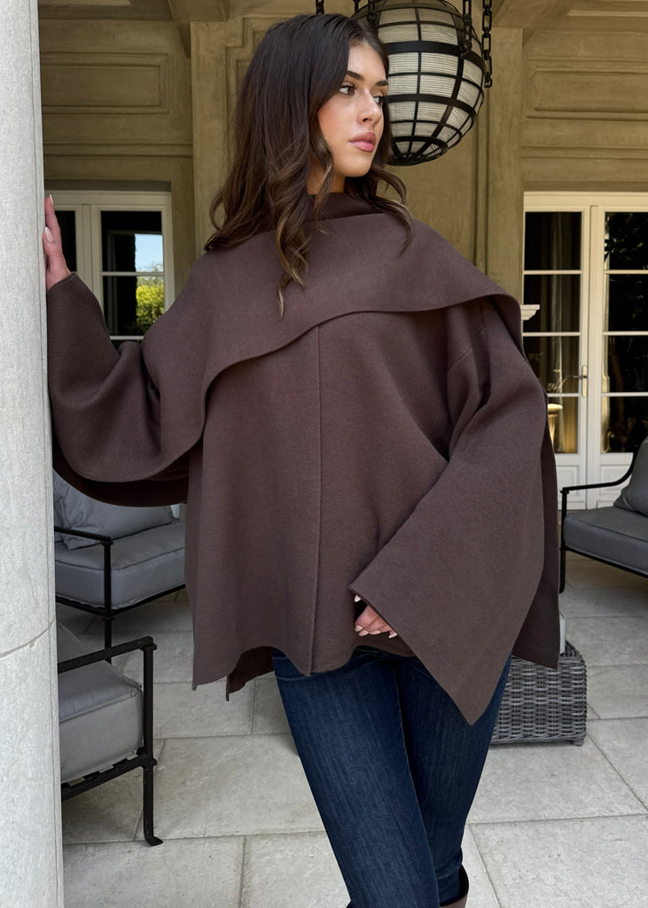 Woman wearing a brown cape in an outdoor setting with a building in the background