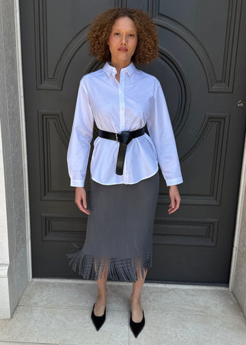 Person wearing a white shirt, black belt, and gray skirt standing in front of a dark door.