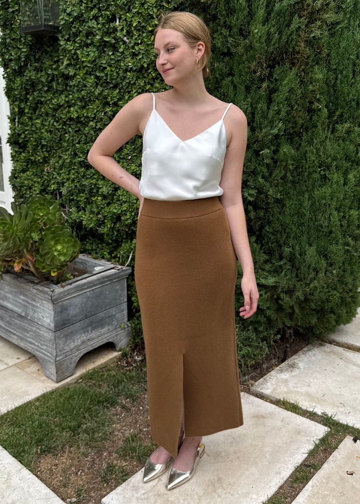 Woman in a white top and brown skirt standing outdoors next to a green bush.