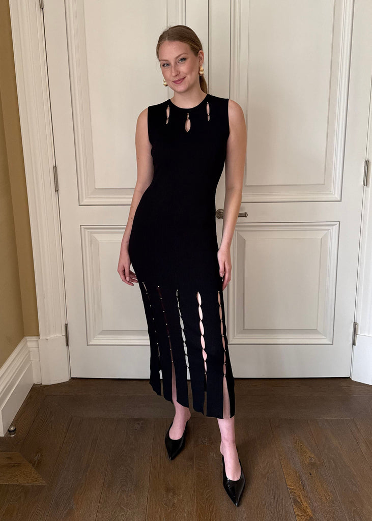 Woman wearing a black sleeveless dress with cut-out details in a room with wooden flooring and white paneled walls.