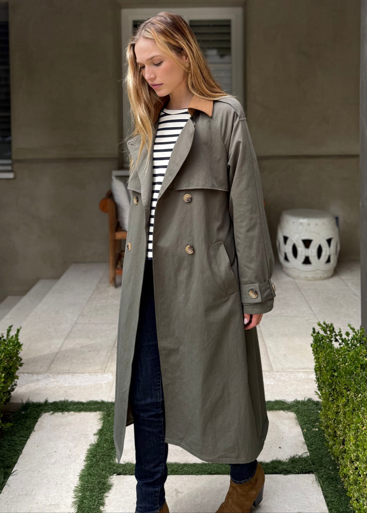 Woman in a trench coat standing in a garden area with a building in the background