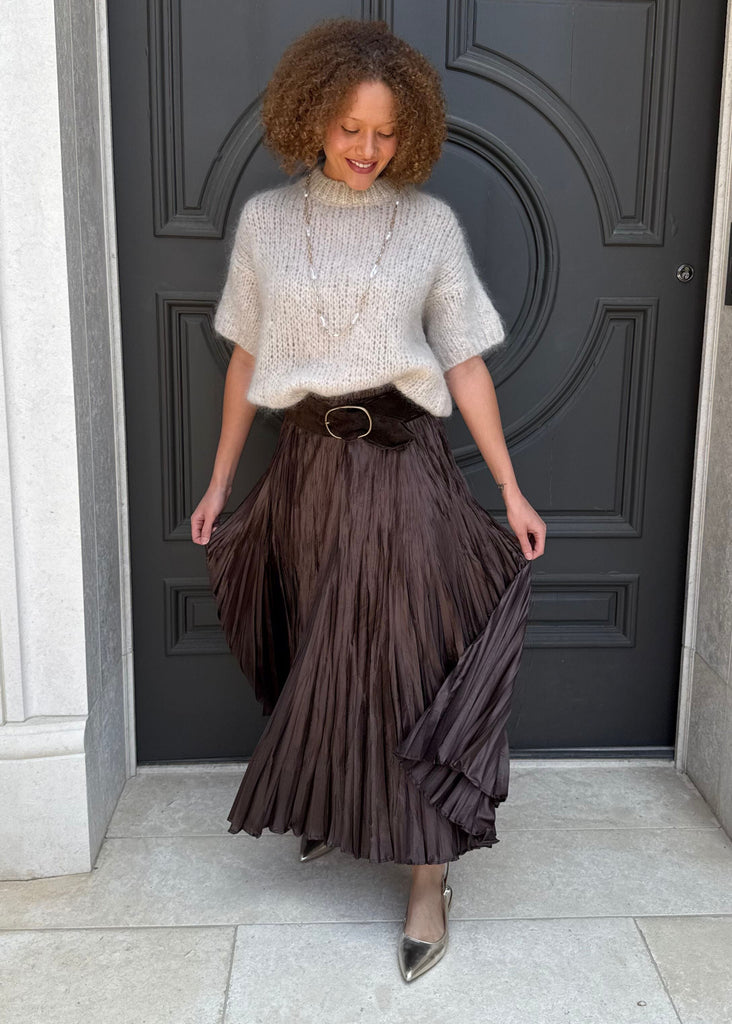 Woman standing in front of a dark gray door wearing a light sweater and brown pleated skirt.