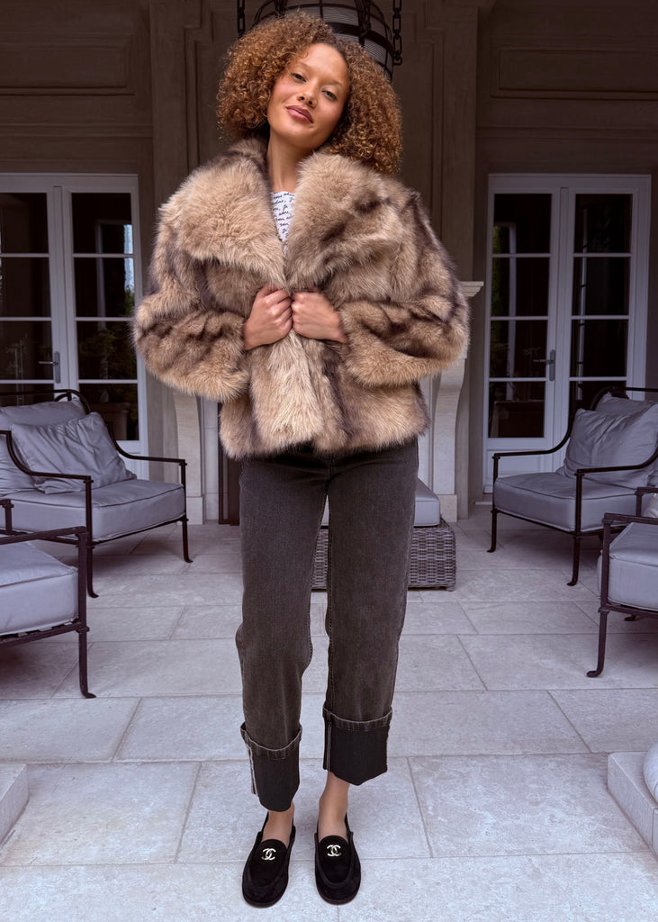 Person wearing a fur coat standing on a patio with chairs and a table in the background.