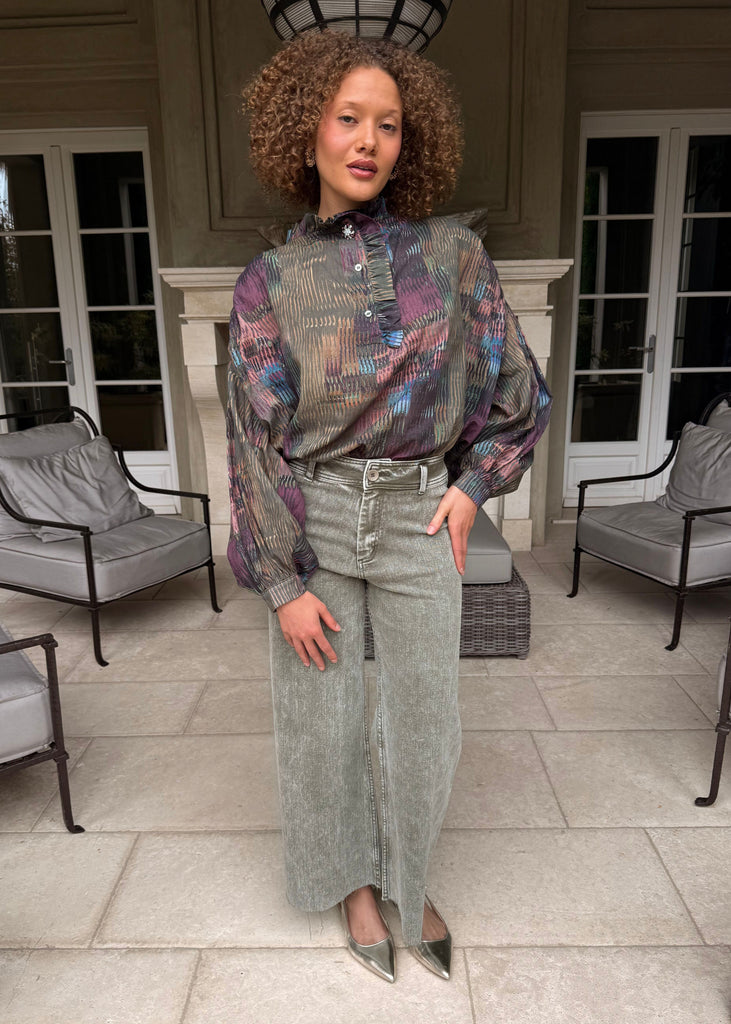 Woman standing in a stylish outfit with a patterned blouse and denim skirt in an outdoor setting.