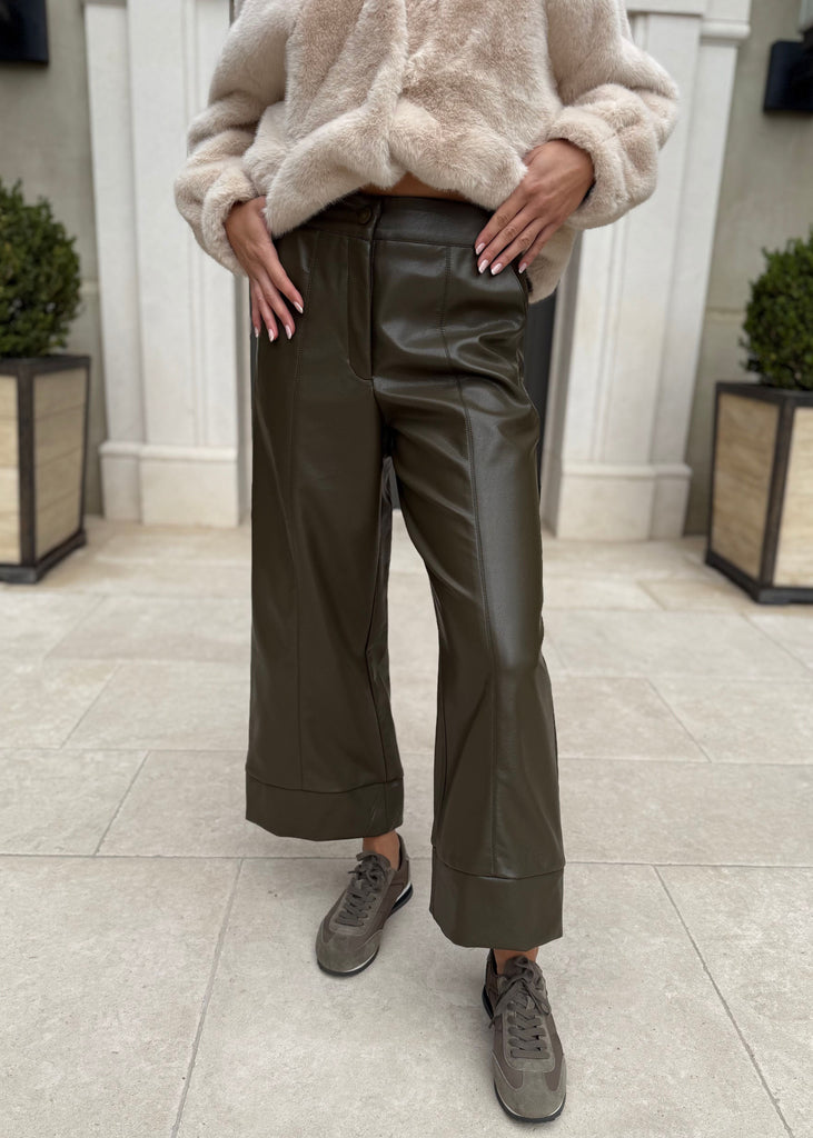 Person wearing a beige sweater and green leather pants standing in a tiled room.