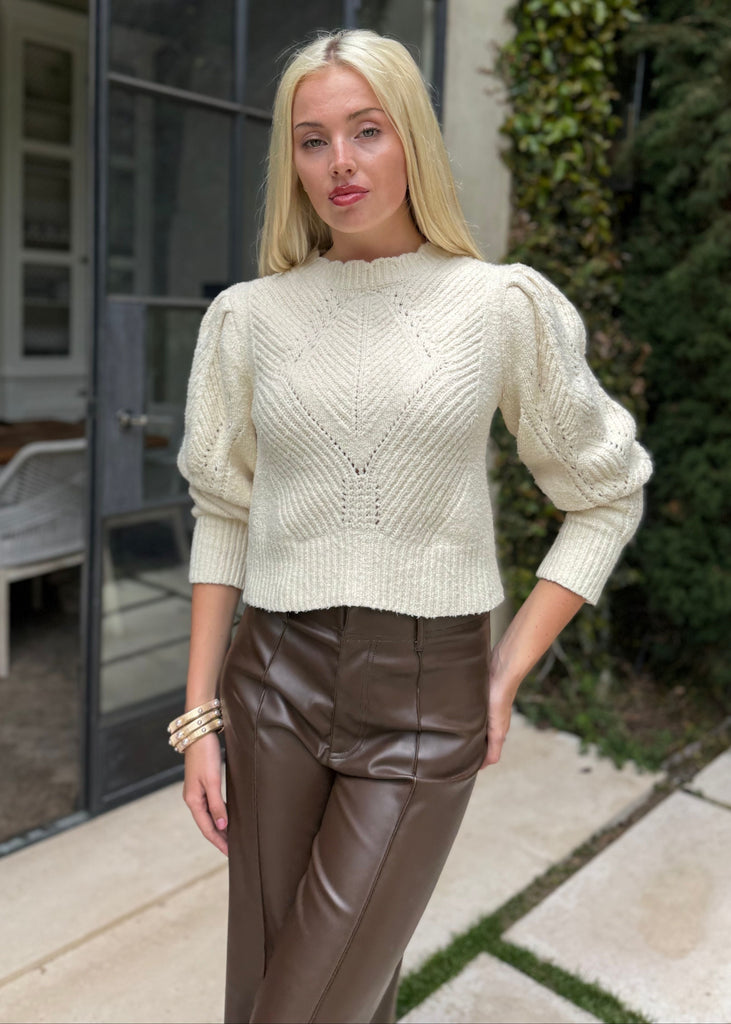 Woman wearing a cream sweater and brown pants standing outdoors.
