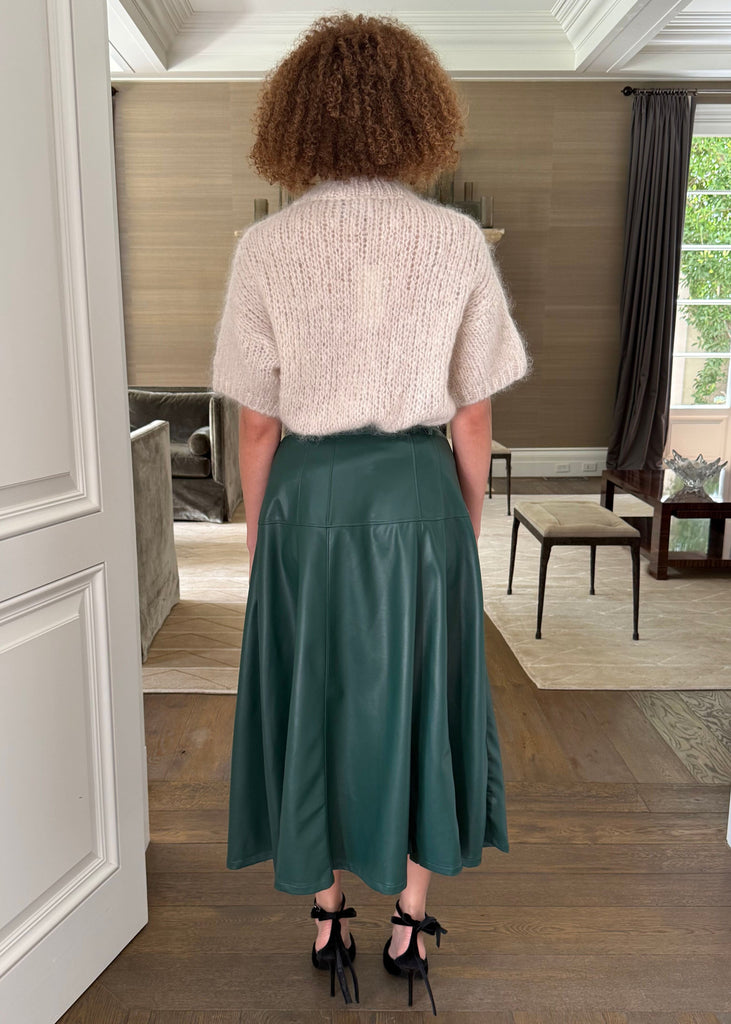 Person wearing a white sweater and green skirt standing in a room with wooden flooring and white walls.