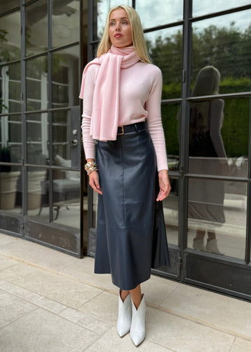 Woman wearing a pink sweater, black leather skirt, and white boots standing in front of a glass building.