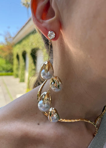 Close-up of a person wearing gold hoop earrings with pearl-like stones outdoors.