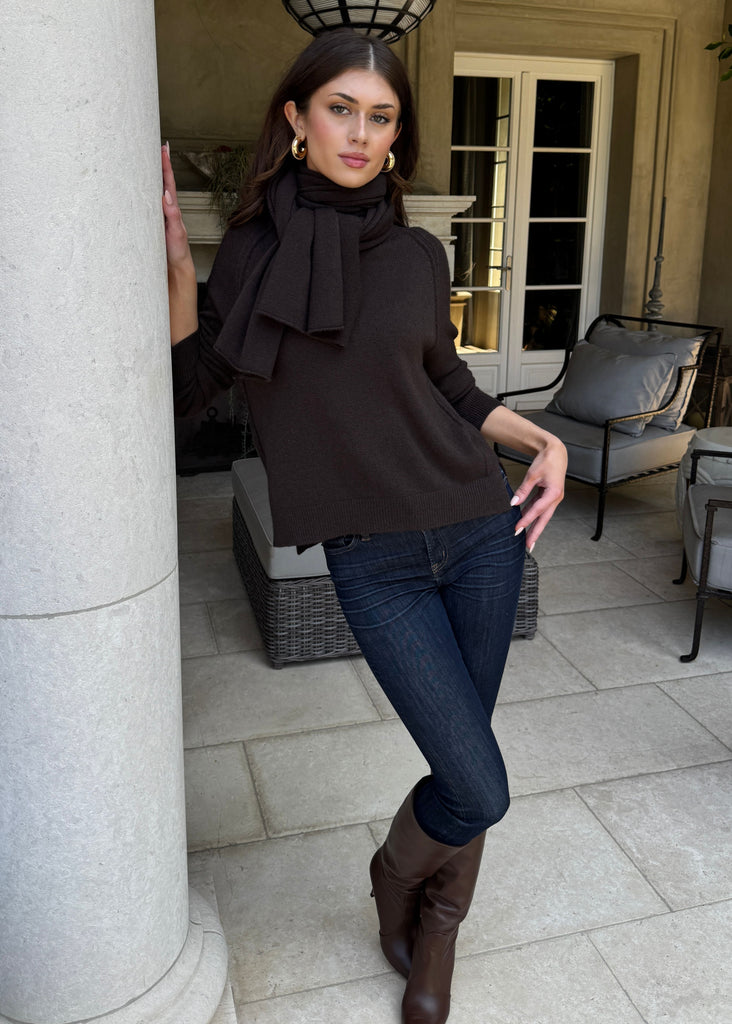 Woman in a brown top and jeans standing in an elegant outdoor setting with columns and furniture.
