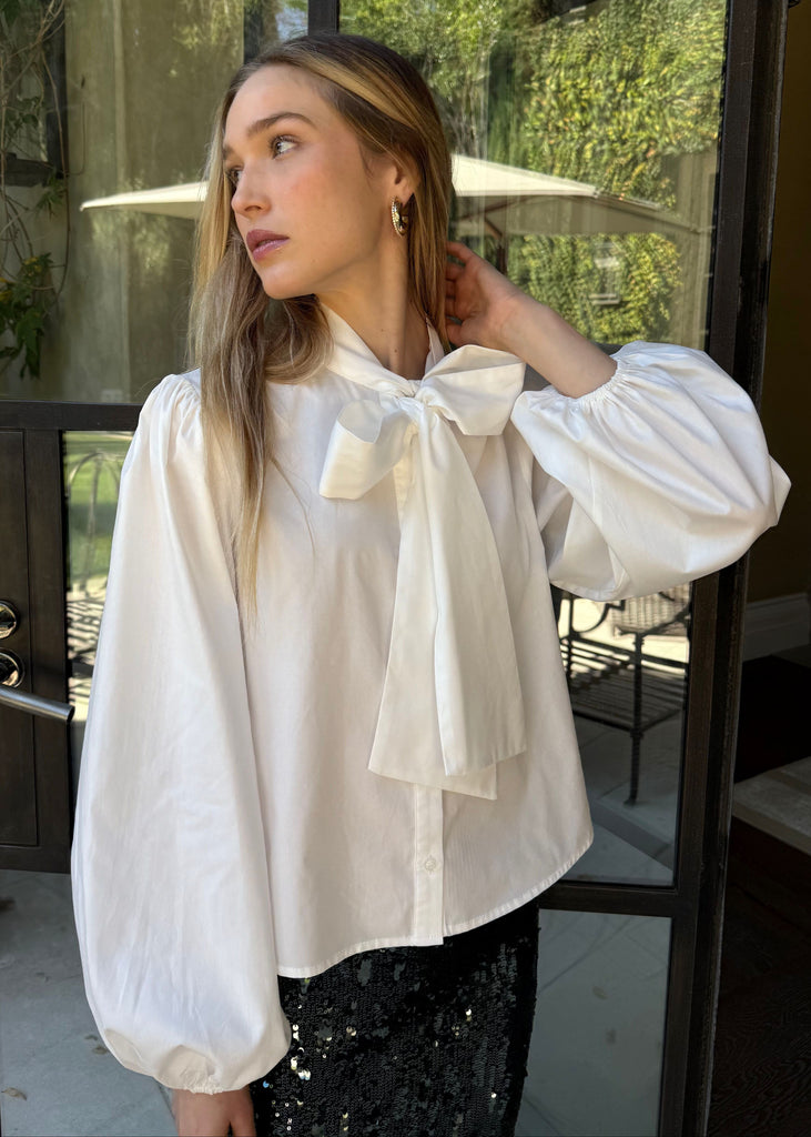 Woman wearing a white blouse with a bow tie in front of a glass door.