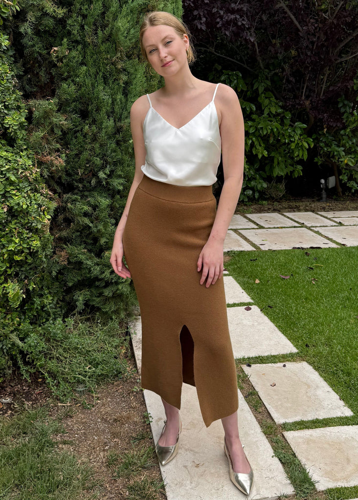 Woman in a white top and brown skirt standing on a garden path with greenery.