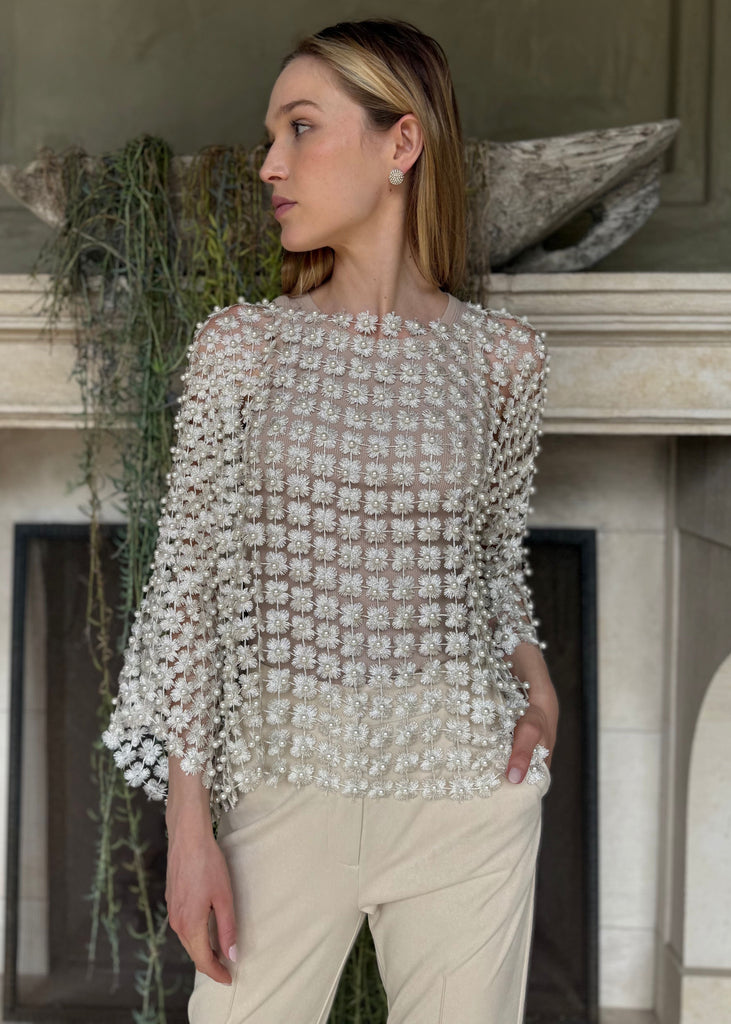 Woman wearing a silver beaded top and white pants standing in front of a fireplace.