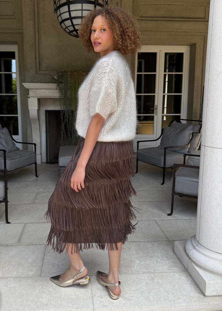 Woman wearing a white sweater and brown fringed skirt standing in an elegant outdoor setting.