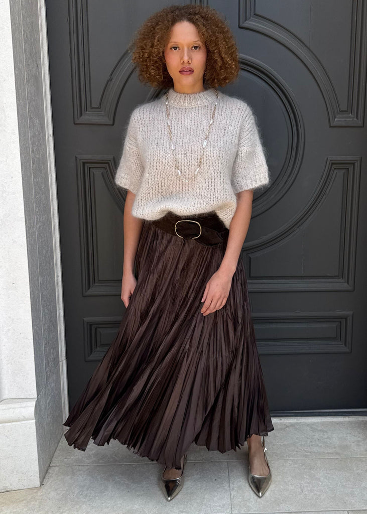 Woman standing in front of a dark gray door wearing a white top and brown pleated skirt.