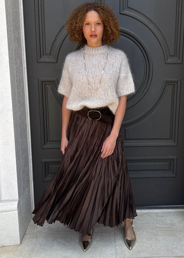Woman standing in front of a dark gray door wearing a white top and brown pleated skirt.