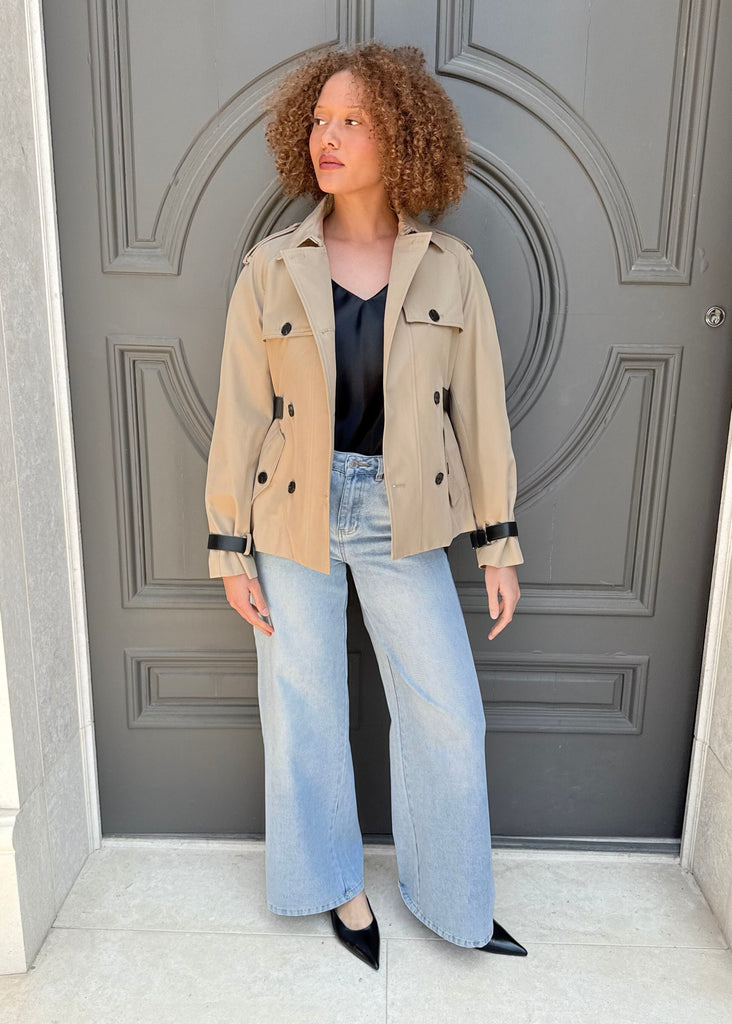 Woman standing in front of a gray door wearing a beige jacket, black top, and light blue jeans.