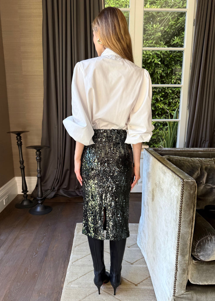 Woman wearing a white blouse and black sequin skirt in a room with a couch and window.