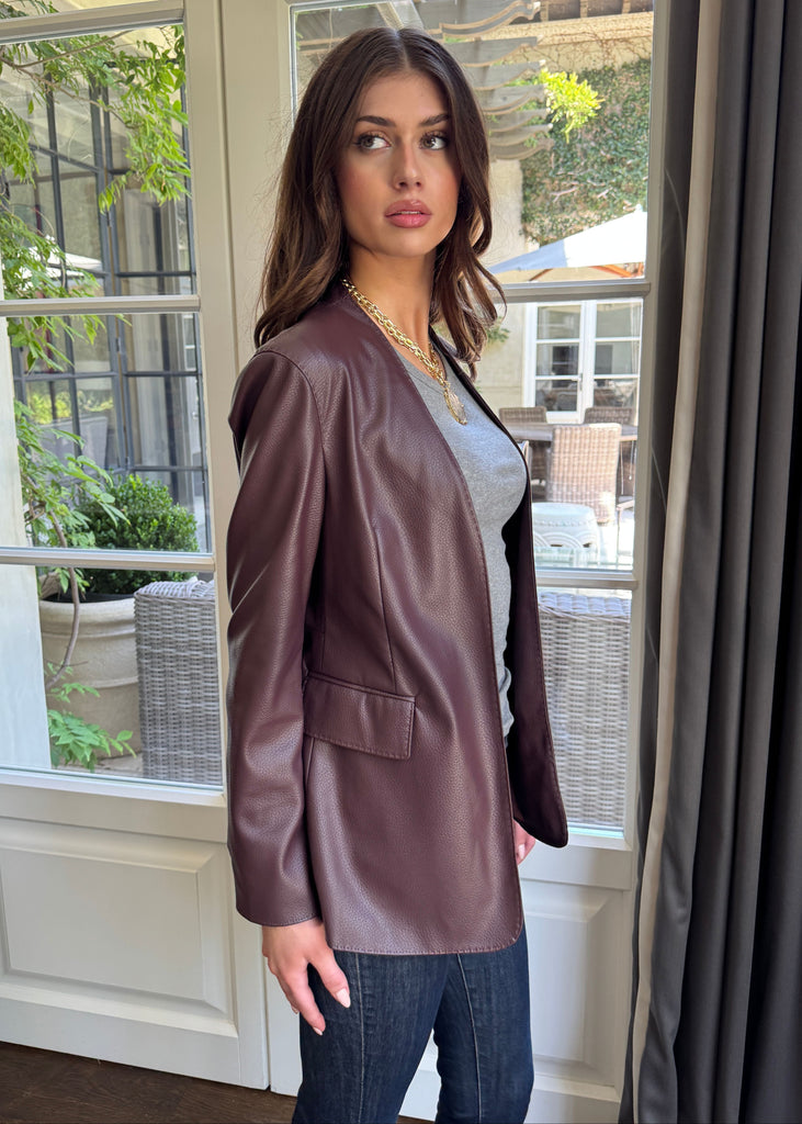 Woman wearing a brown leather jacket standing in front of a window with a view of greenery outside.