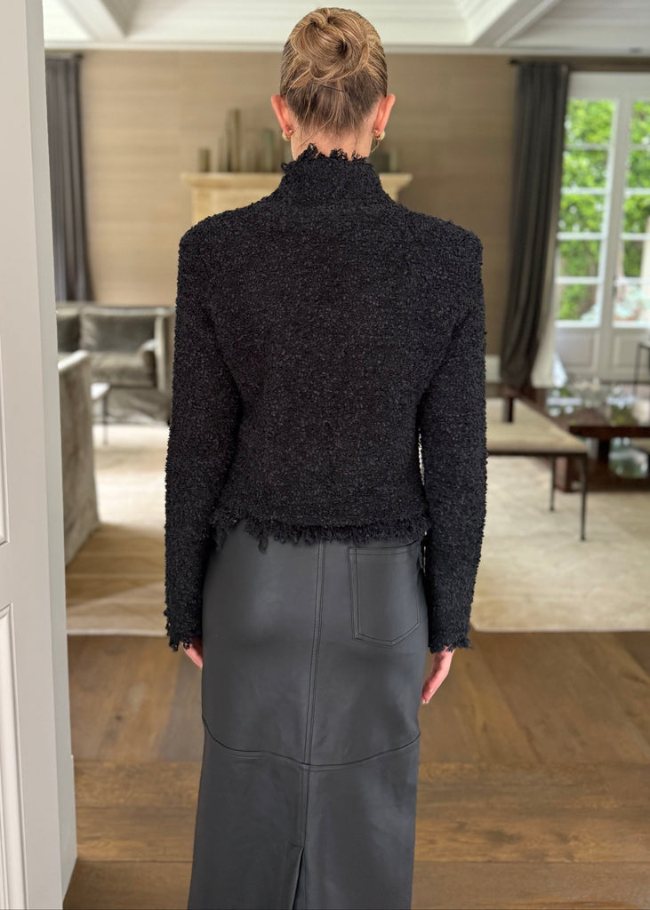 Person wearing a black textured top and gray pants standing in a room with wooden flooring and white walls.