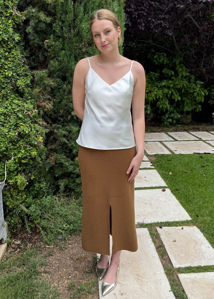 Woman in a white top and brown skirt standing on a garden path with greenery.