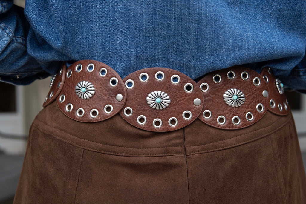 Brown leather belt with decorative conchos worn by a person in a blue shirt and brown pants.