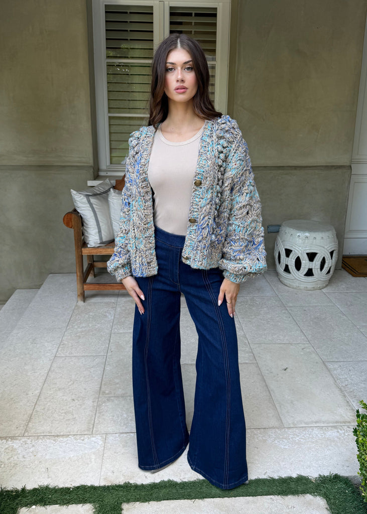 Woman standing outdoors in front of a house wearing a patterned cardigan, beige top, and blue flared pants.